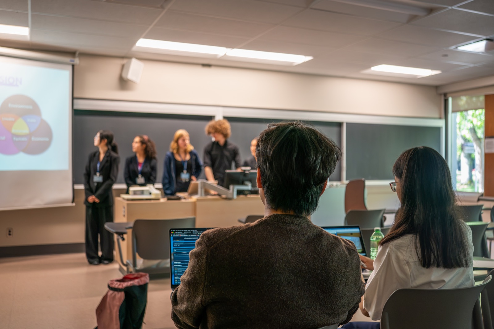 A team presenting to the room while other students watch from their laptops